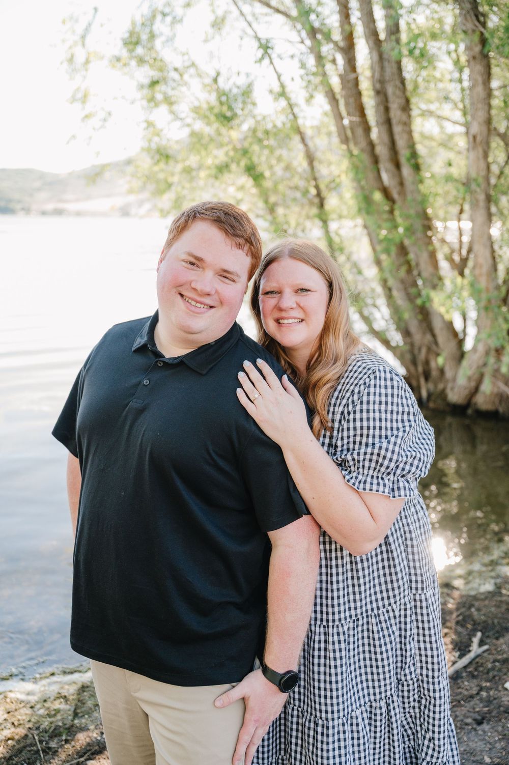 Smiling couple posing outdoors near a lake with trees, man in black polo and woman in gingham dress.