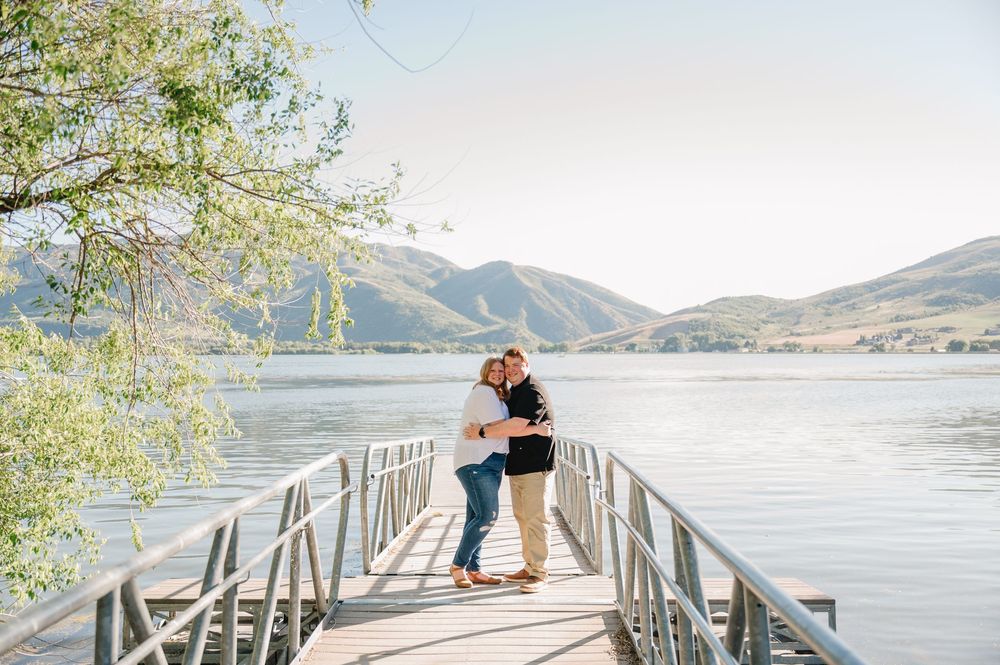 Couple embracing on a wooden dock overlooking a serene mountain lake under clear blue skies.