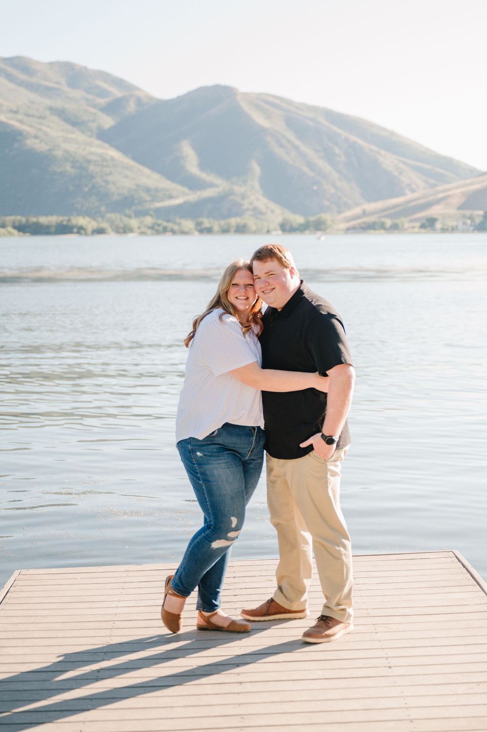 Couple embracing and smiling on a wooden dock with a scenic mountain lake backdrop on a sunny day.
