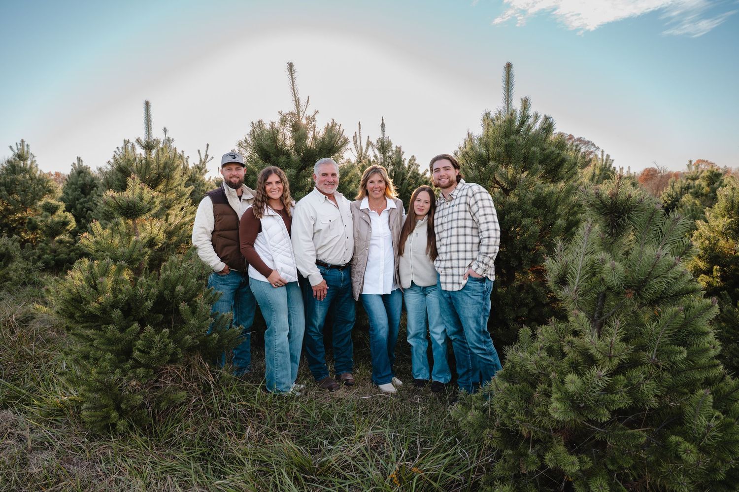 Family of six posing among pine trees at a Christmas tree farm with a rainbow in the background.
