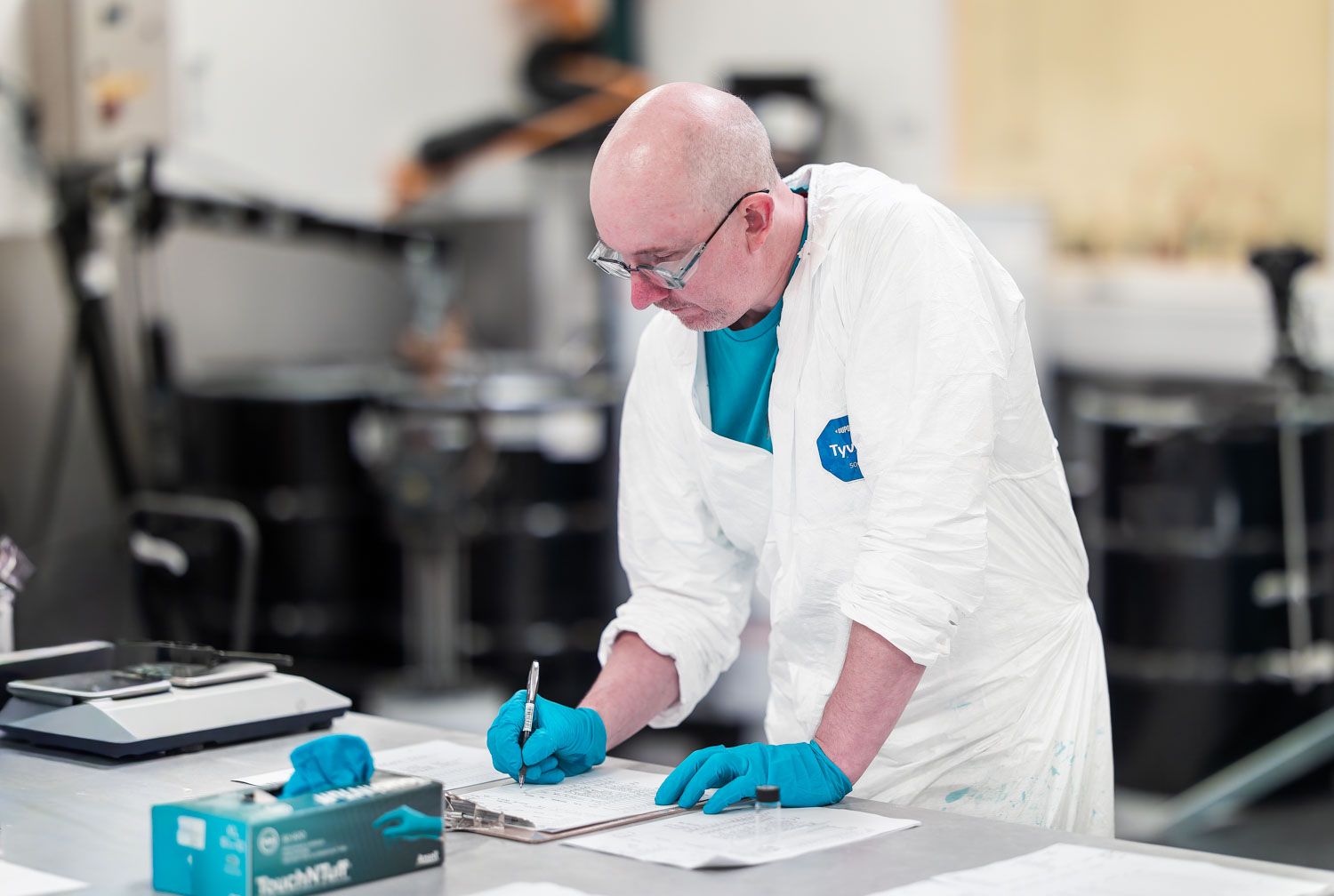 Factory inspector in white lab coat and blue gloves writing notes on clipboard in a laboratory setting.