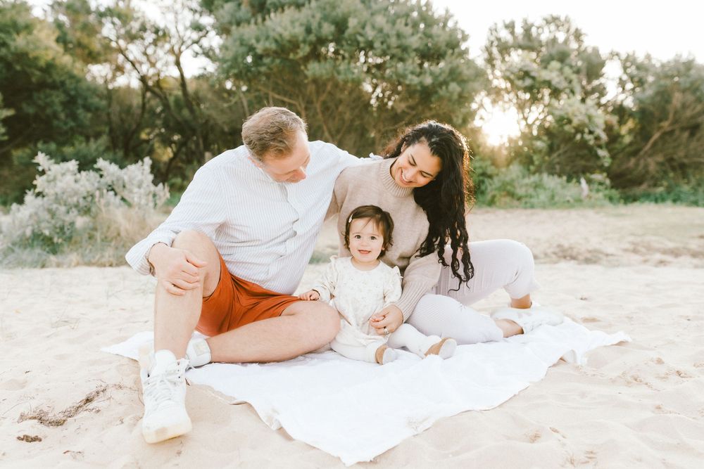 Family of three sitting on white blanket on sandy beach, parents smiling at toddler in golden sunset light.