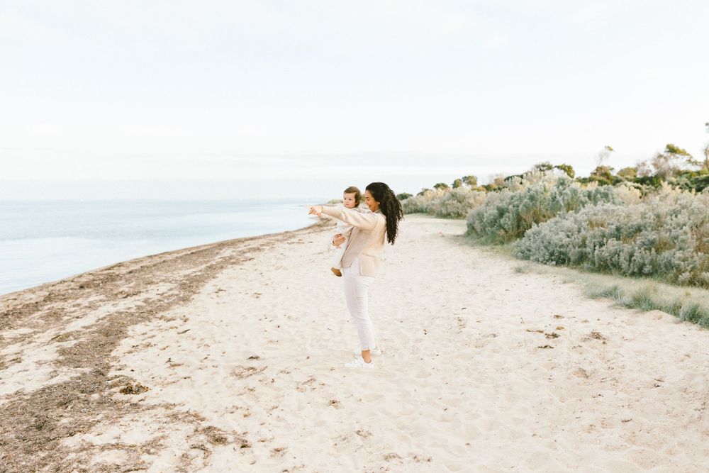 A woman in white outfit playfully poses on a sandy beach path with ocean views and coastal shrubs.