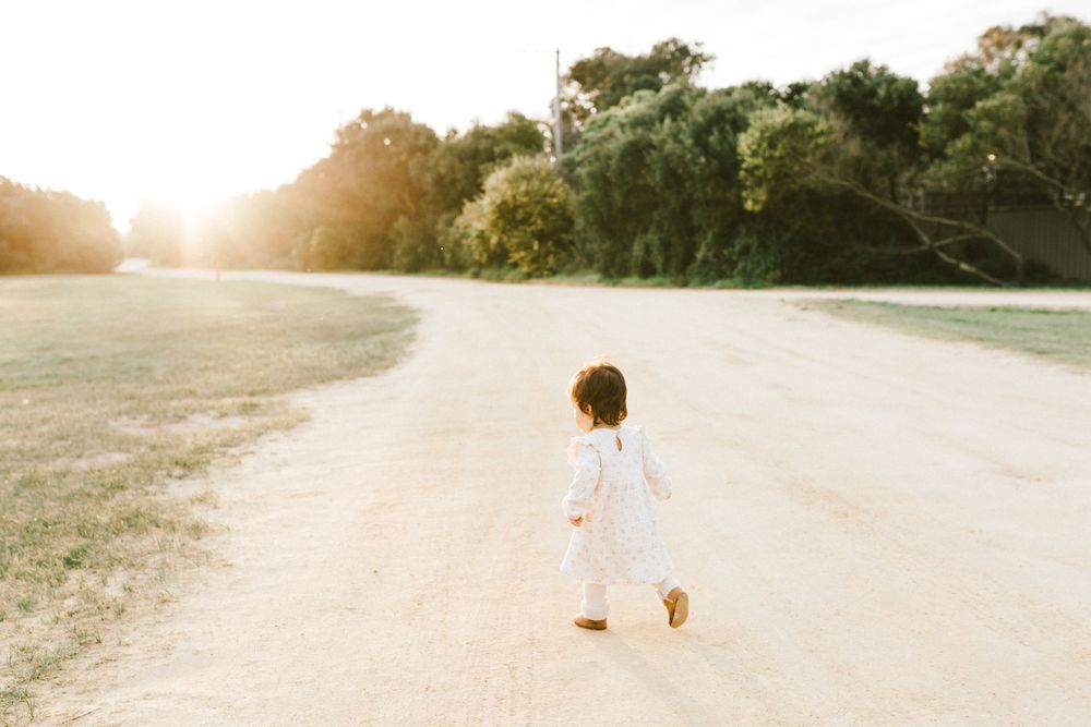 A toddler in a white dress walks along a sunny dirt path surrounded by lush green trees at golden hour.