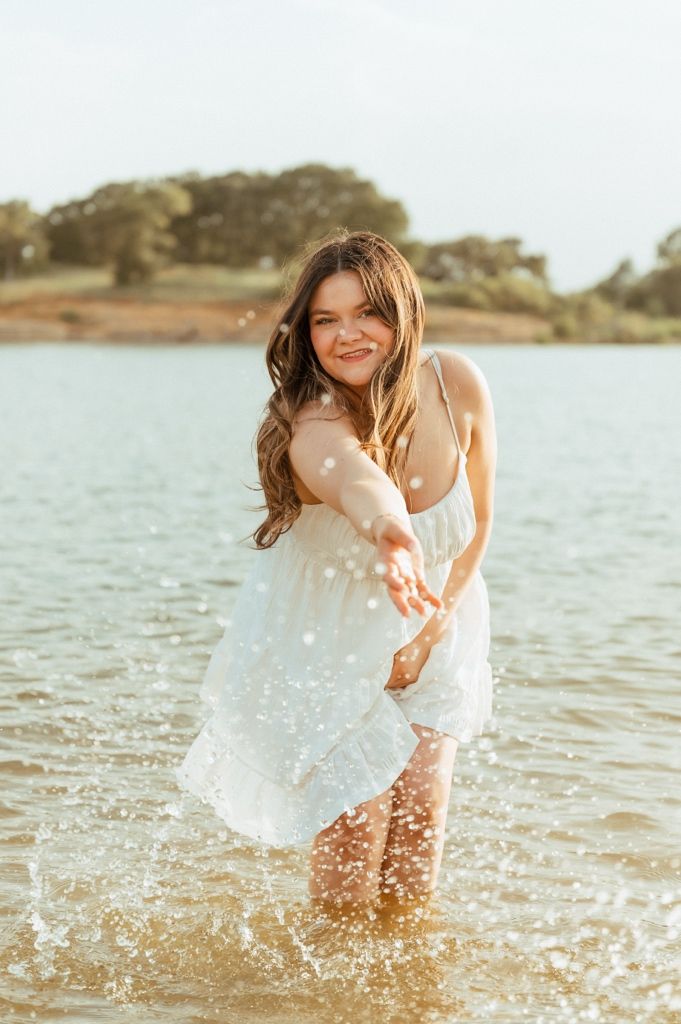 Young woman in white dress splashing water in a lake, smiling joyfully on a sunny day.