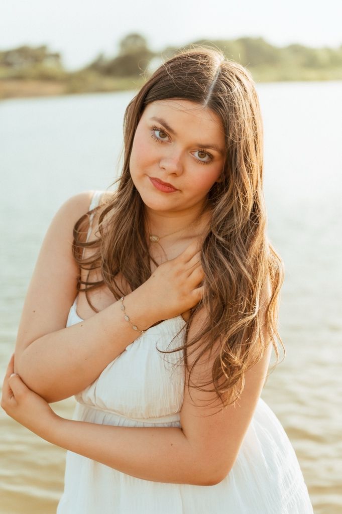 Young woman in white dress posing by a lake, with long brown hair and a delicate gold necklace.
