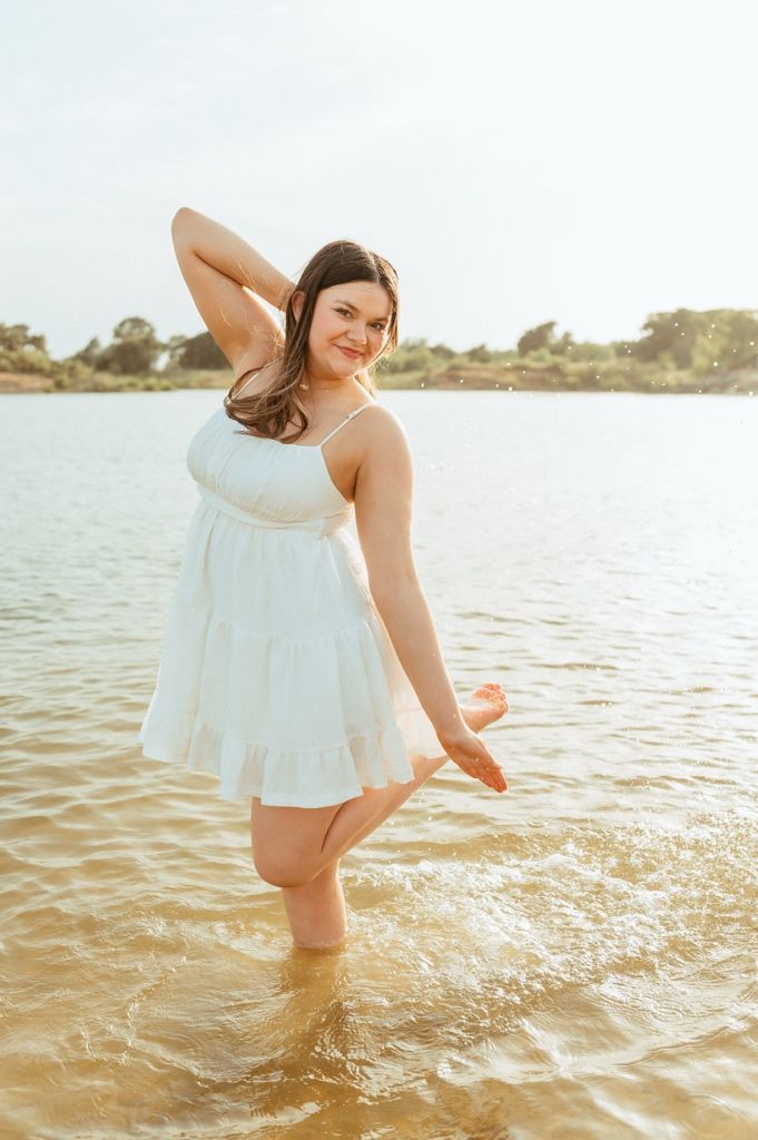 Young woman in white sundress wading in shallow lake water, smiling over shoulder in golden sunlight.