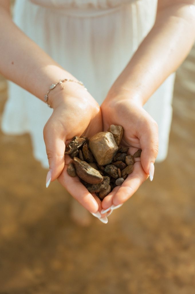 A woman in a white dress holds a handful of small golden-brown river rocks in her cupped hands, wearing a delicate bracelet.