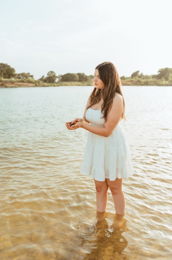 Young woman in white sundress standing in shallow lake water, holding something small, with trees and sky in background.