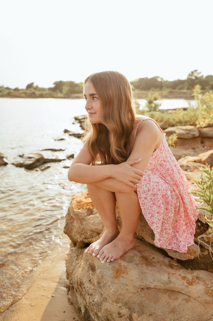 Barefoot teenage girl in floral pink dress sitting on rocks by a lake, gazing into the distance.