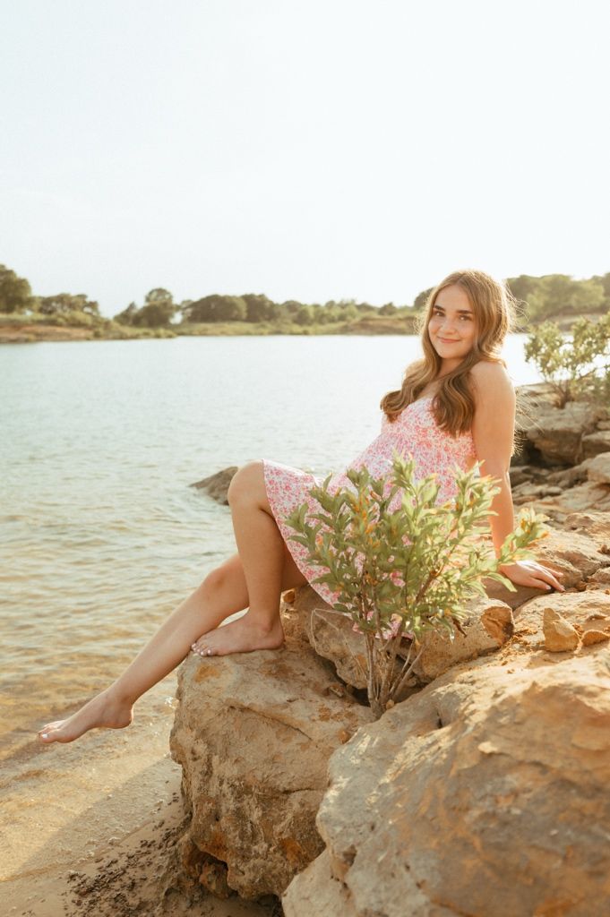 Young woman in pink floral dress sitting on rocks by a serene lake, smiling, with a small green plant nearby.