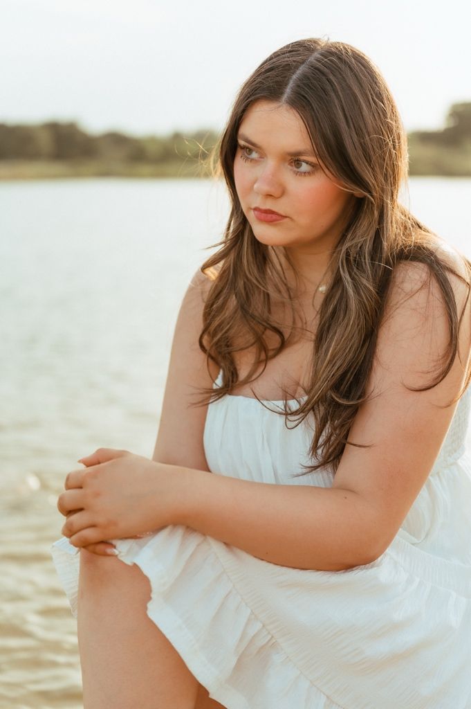 Young woman in white dress sitting by a lake at golden hour, looking thoughtfully to the side.