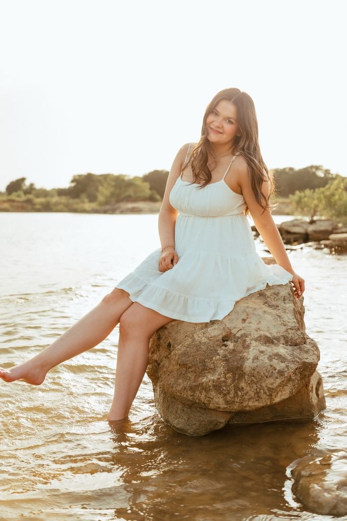 Young woman in a light blue sundress sitting on a rock in shallow water, smiling at the camera on a sunny day.