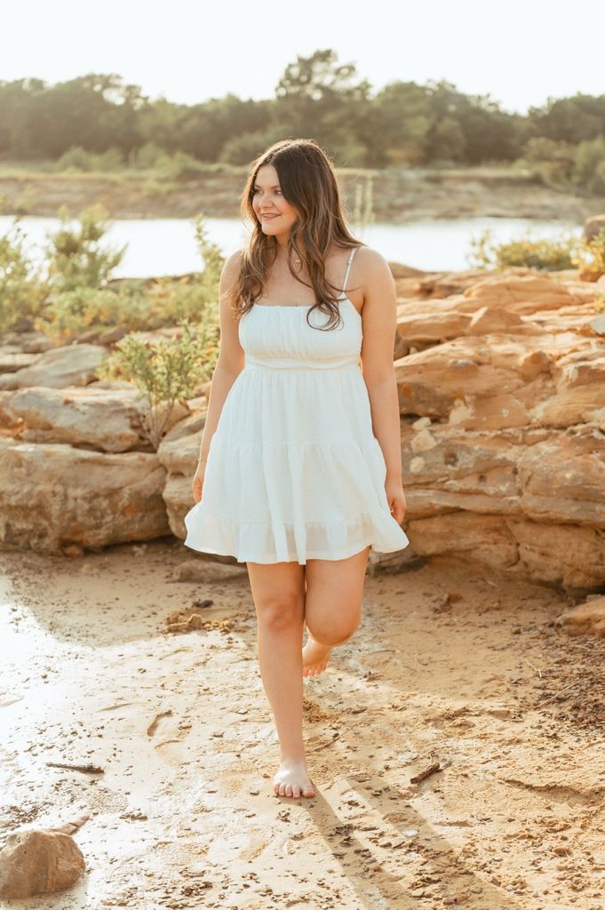 Young woman in white sundress walking barefoot on sandy rocky terrain near water at golden hour.