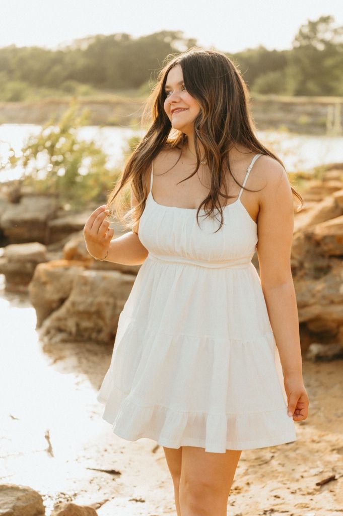 Young woman in white sundress posing near rocky shoreline in golden hour light.