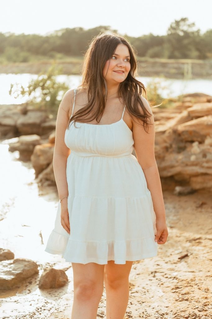 Young woman in white sundress standing near rocky shoreline at golden hour sunset.