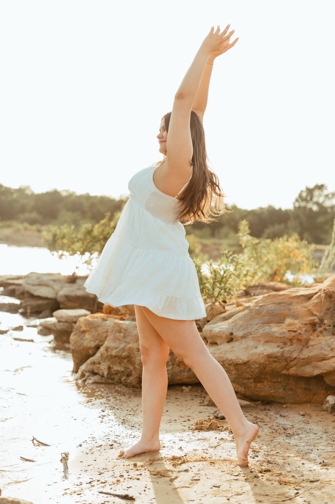 Woman in white dress dancing barefoot on rocky shore with arms raised, bathed in golden sunlight.
