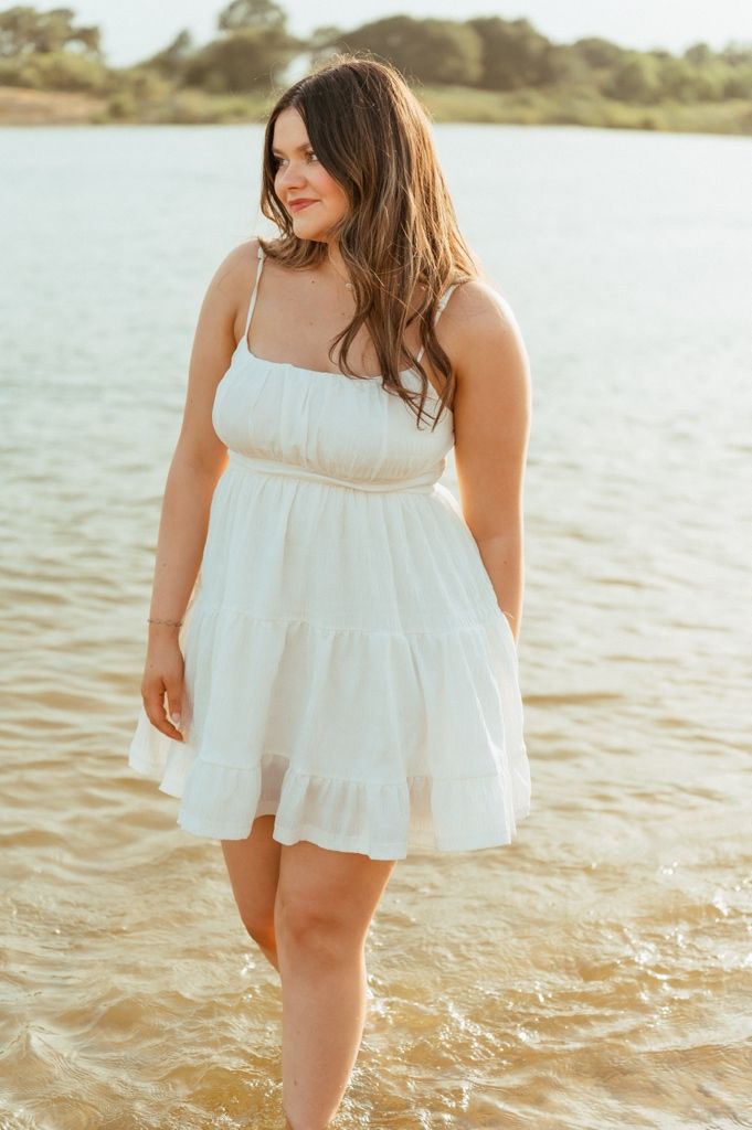 Young woman in white tiered sundress standing in shallow water near shore on a sunny day.
