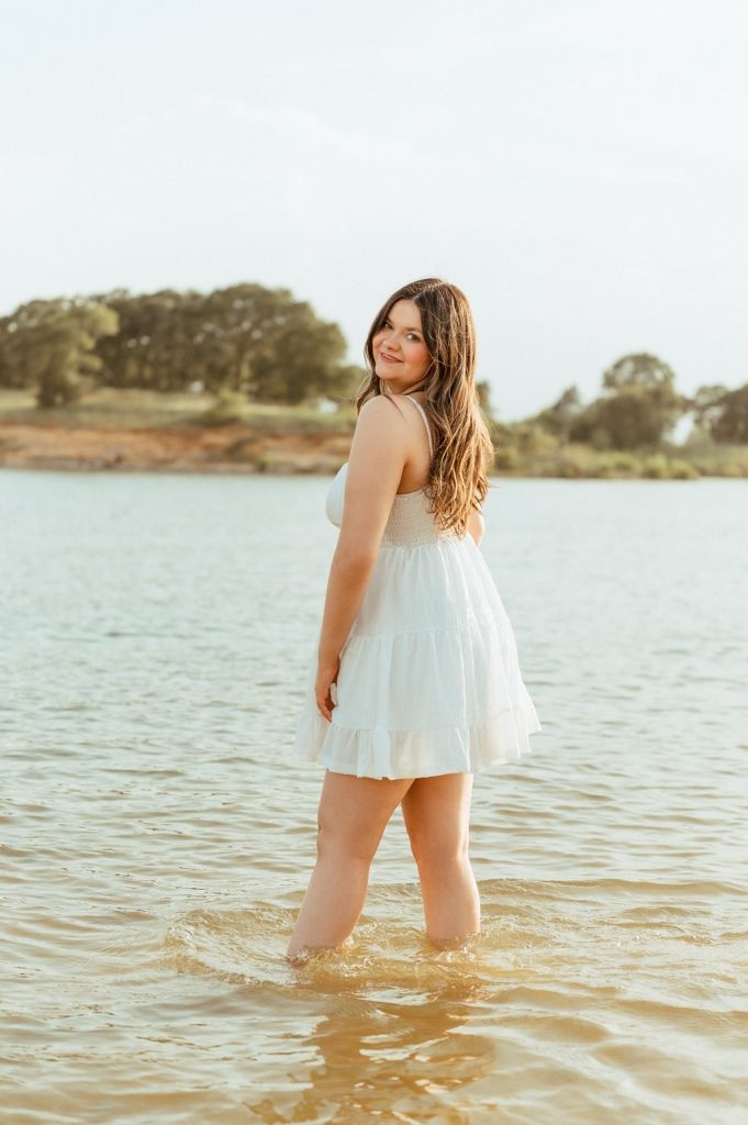 Young woman in white sundress standing in shallow lake water, looking back over shoulder with trees in background.