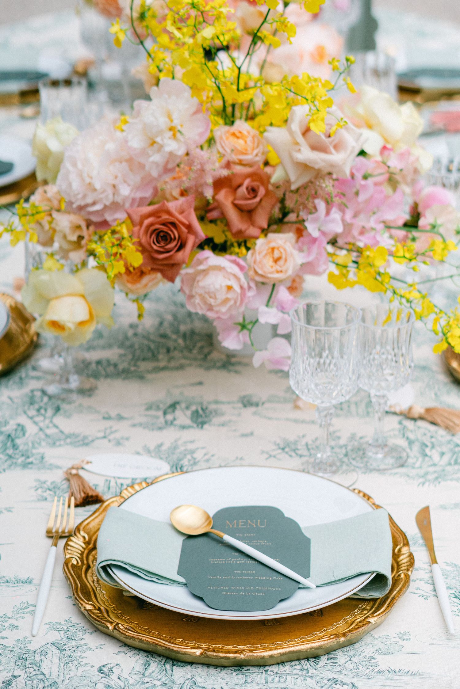 Elegant wedding table setting with gold charger, sage menu card, and lush floral centerpiece of pink, peach roses and yellow blooms.