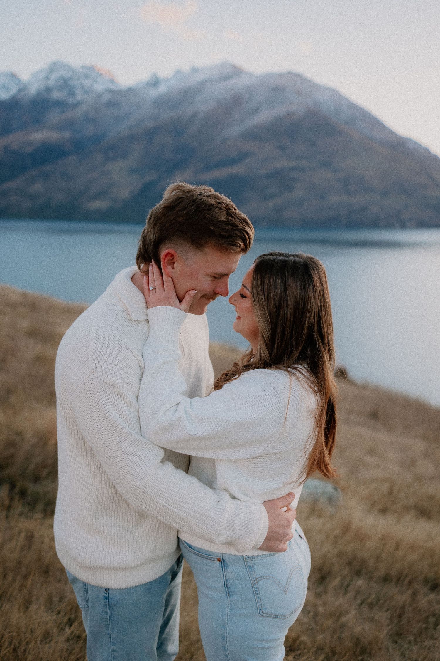 Couple in white sweaters and jeans embracing near a lake with snow-capped mountains in the background.