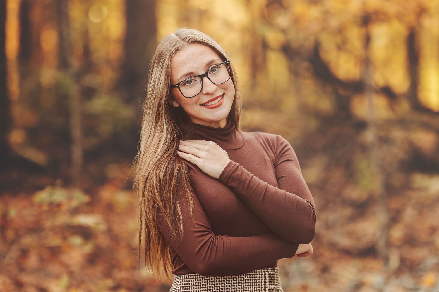 A young blonde woman in glasses, brown turtleneck, and knit skirt poses on a rural path surrounded by autumn foliage.
