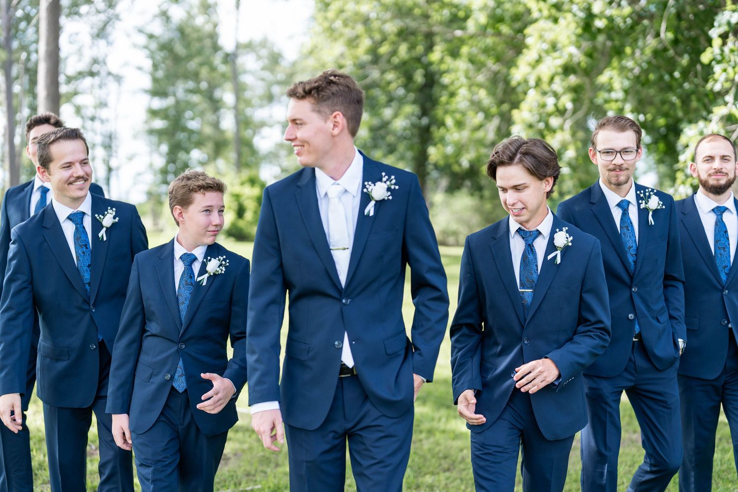 Groomsmen in navy blue suits with boutonnieres walk outdoors on grass with trees in background at a wedding.