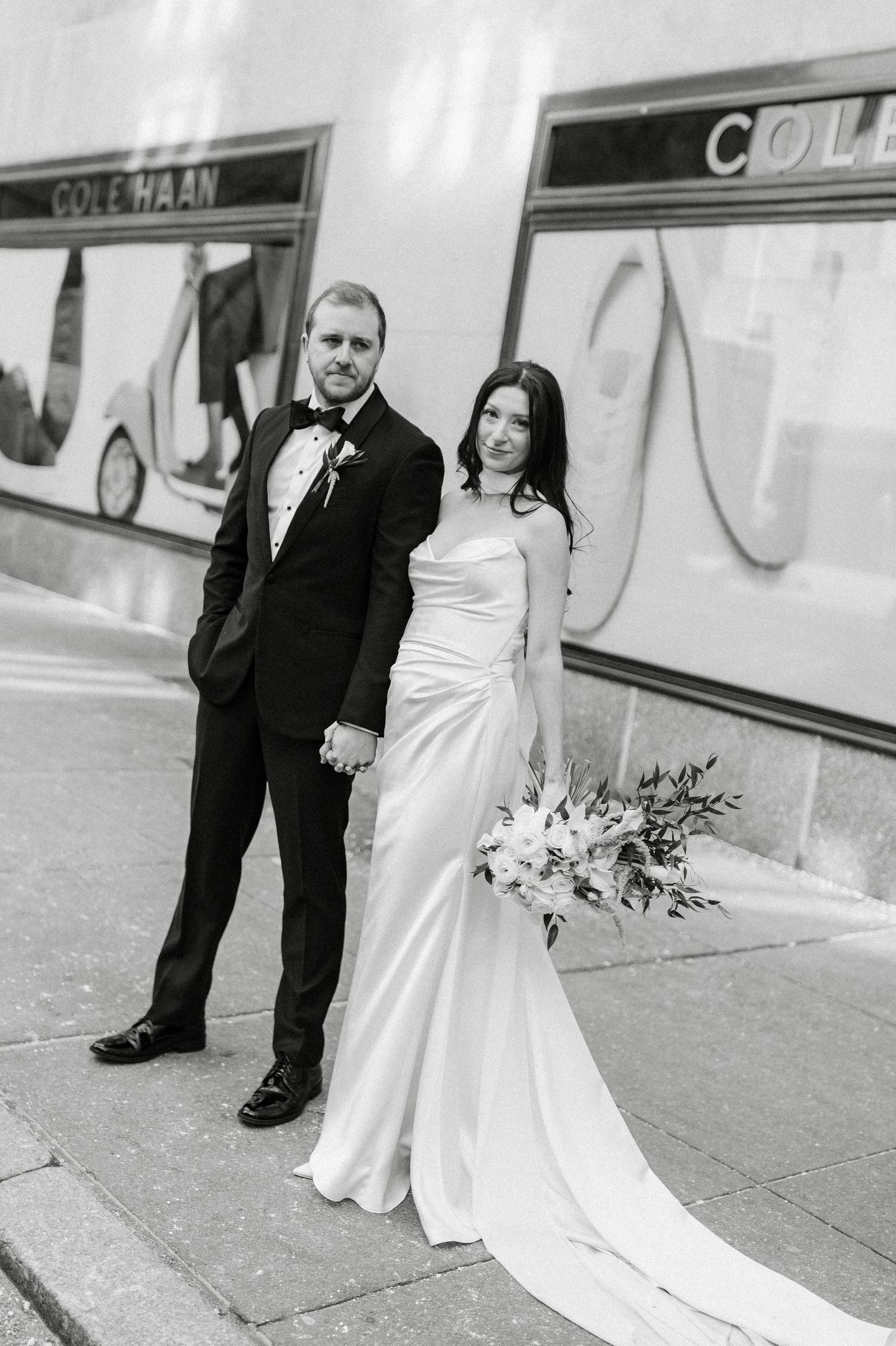 Black and white photo of a bride and groom holding hands outside a Cole Haan store, bride holding a floral bouquet.