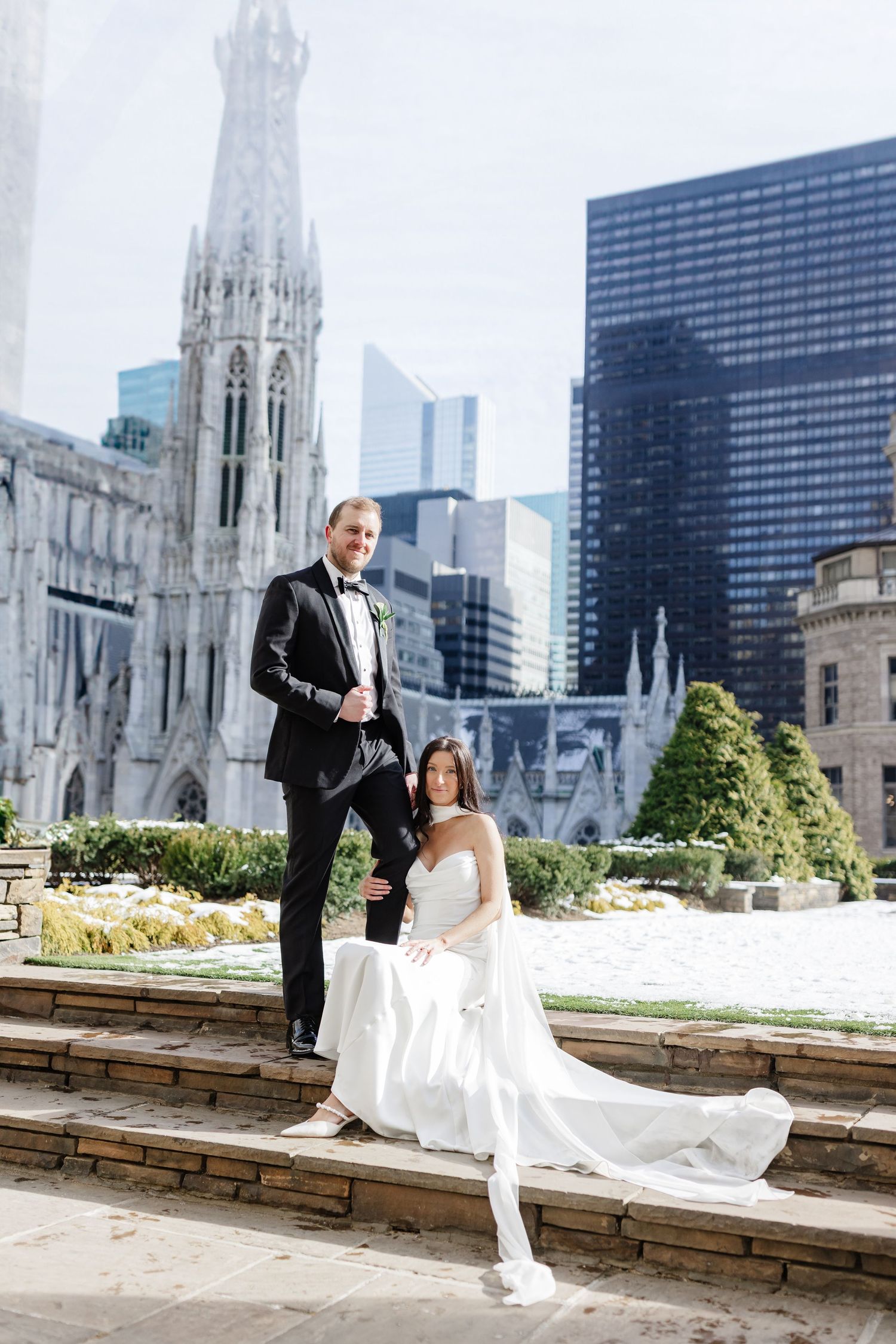 Bride in white gown and groom in black tuxedo pose on stone steps with NYC skyline and Gothic church backdrop.