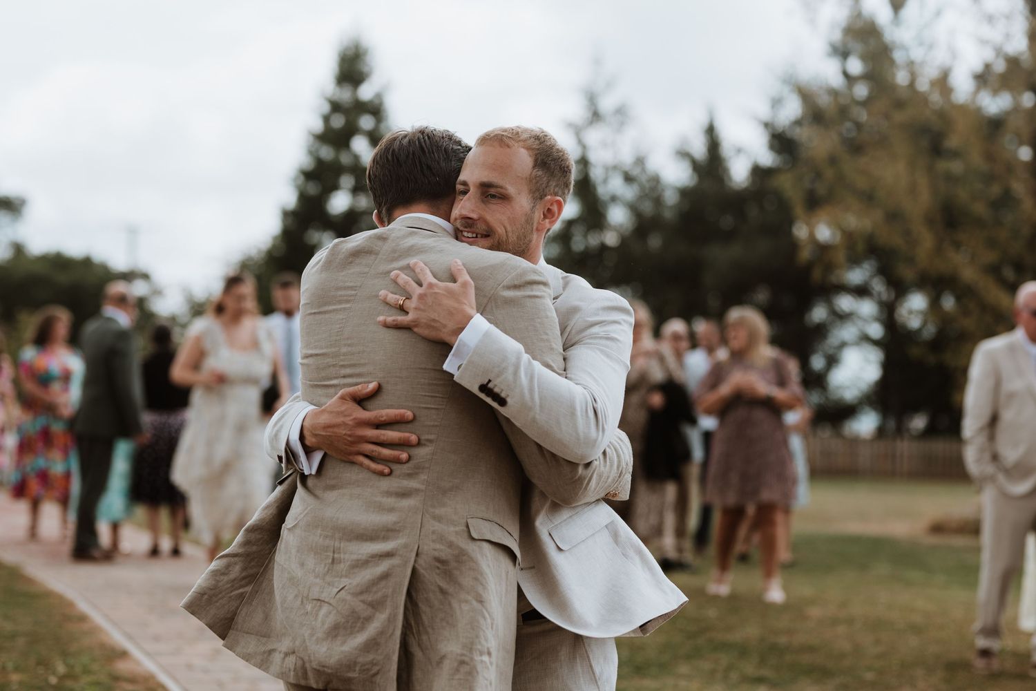 Groom and guest share a heartfelt hug outdoors at a wedding ceremony with guests gathered in the background.