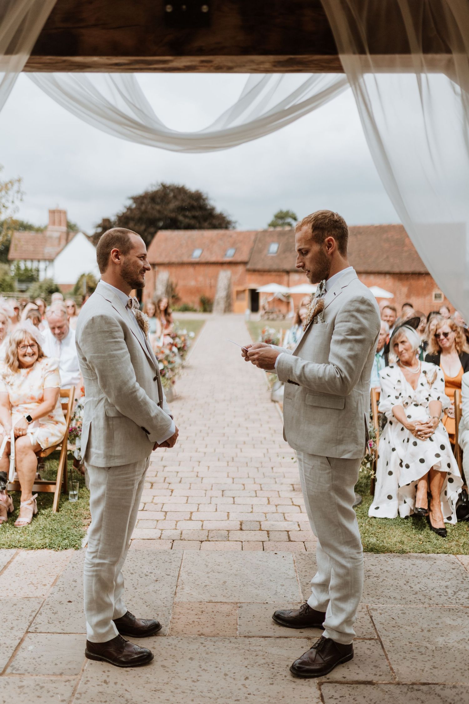 Two grooms in matching light grey suits exchange vows at an outdoor wedding ceremony with guests seated behind them at Moat Hall Barns