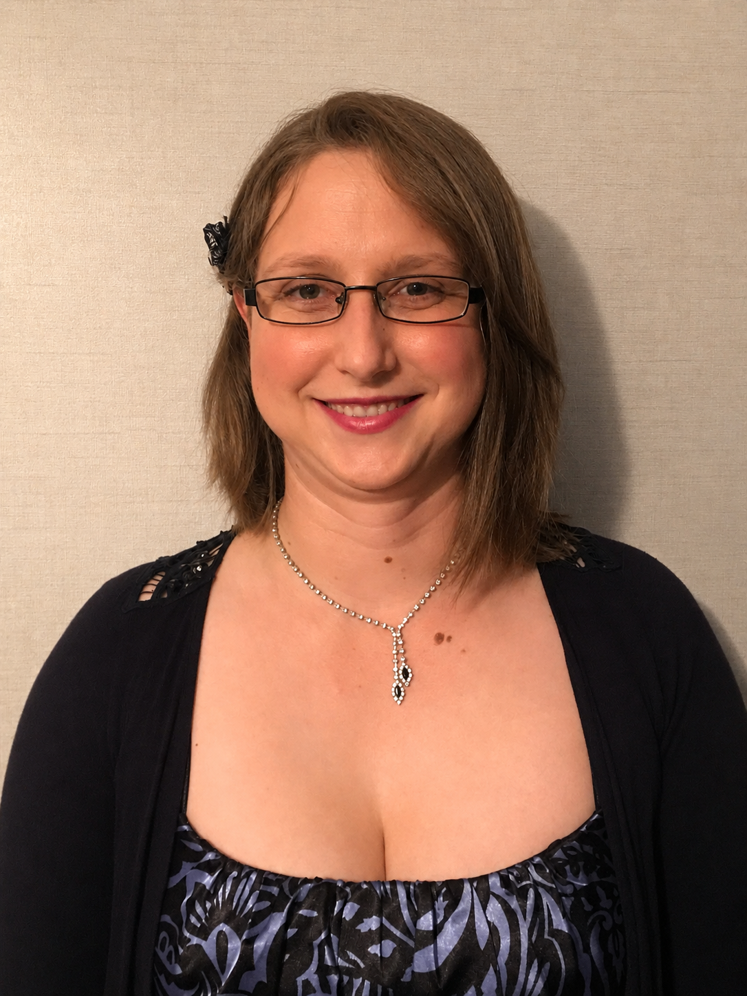 Woman with glasses and brown hair wearing a navy cardigan and floral top with a silver necklace, smiling indoors.
