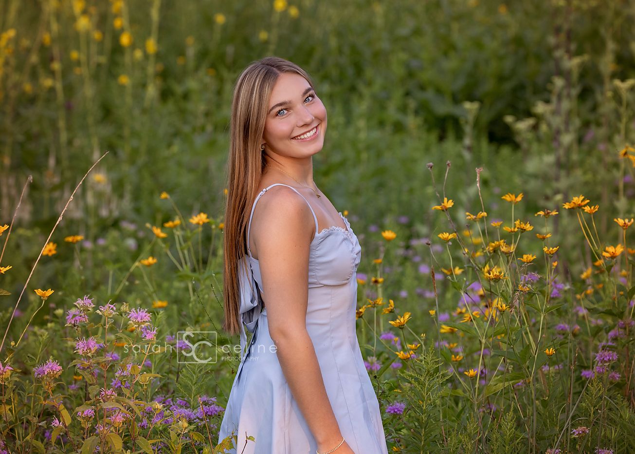 A smiling teenage girl in a light blue dress poses among wildflowers and tall grasses in a sunny outdoor field.