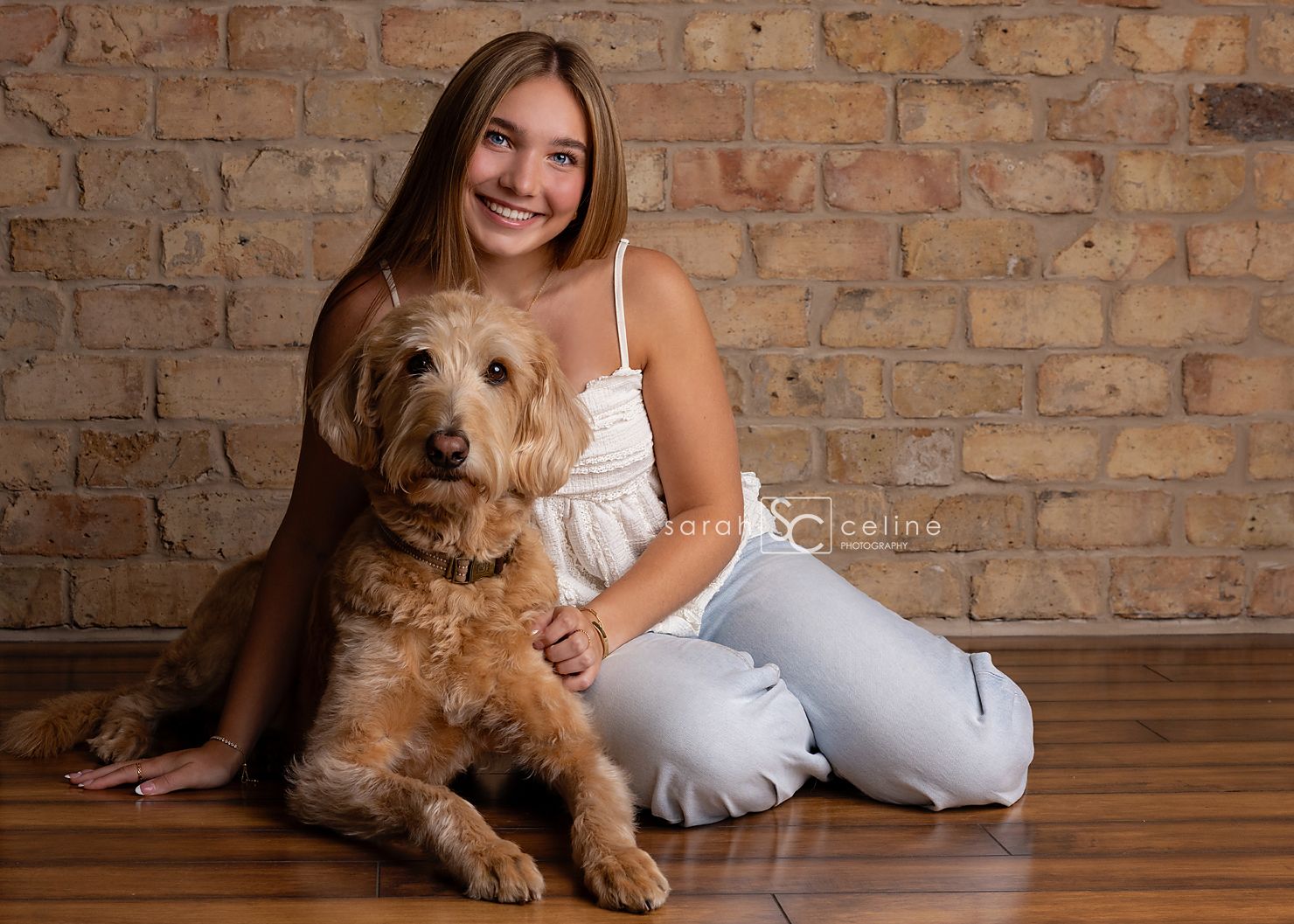 Smiling teenage girl in white top and jeans poses with a fluffy golden doodle dog against a brick wall backdrop.