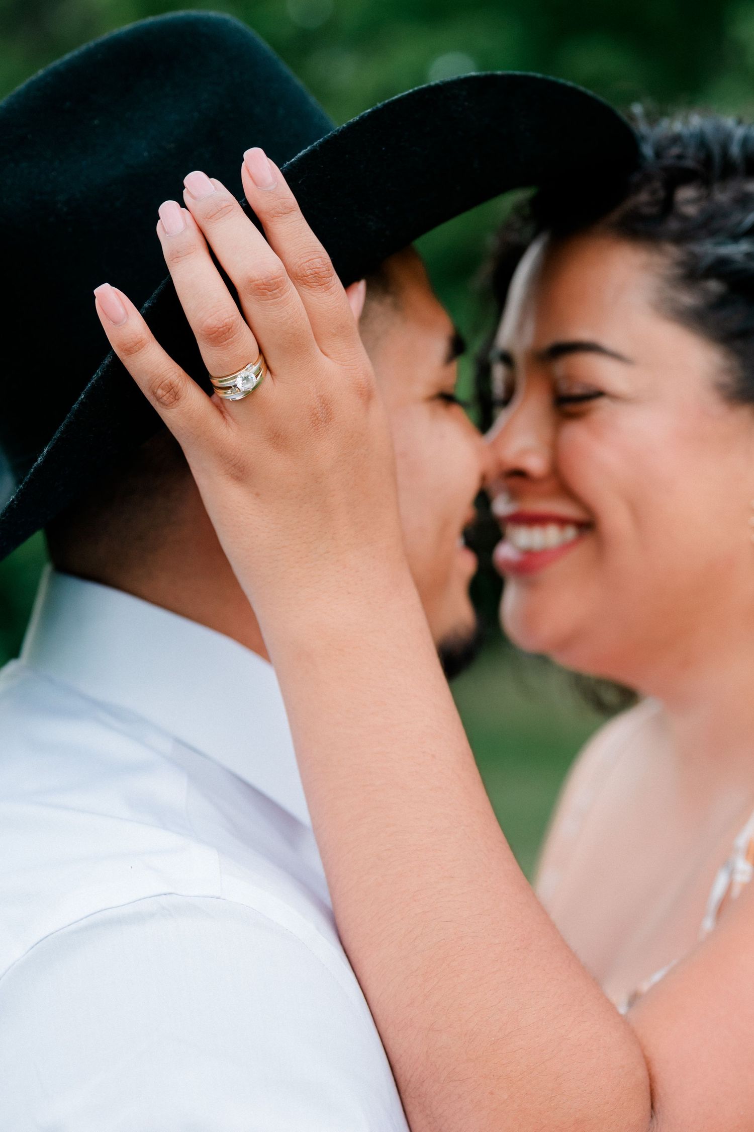 Two women sharing a romantic moment, one wearing a black hat and engagement ring, smiling closely outdoors.