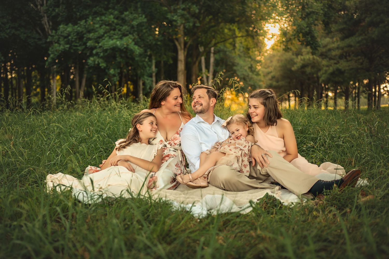 Family of five sitting on blanket in lush green meadow, laughing together during golden hour sunset.
