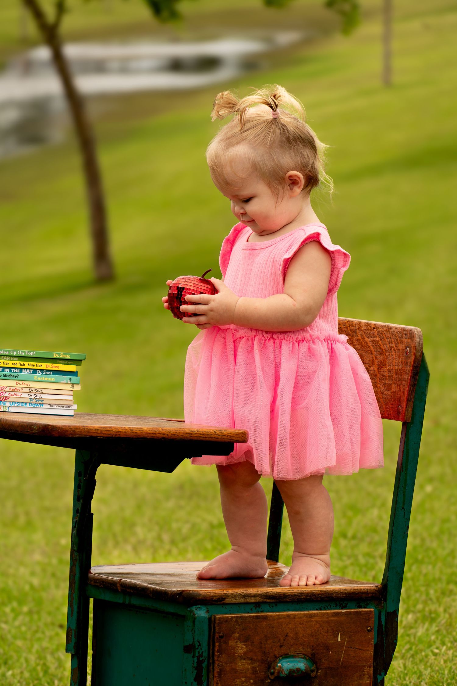 Toddler holds and looks at an apple being held by her in hand. Toddler is standing on a vintage desk chair outside in a park