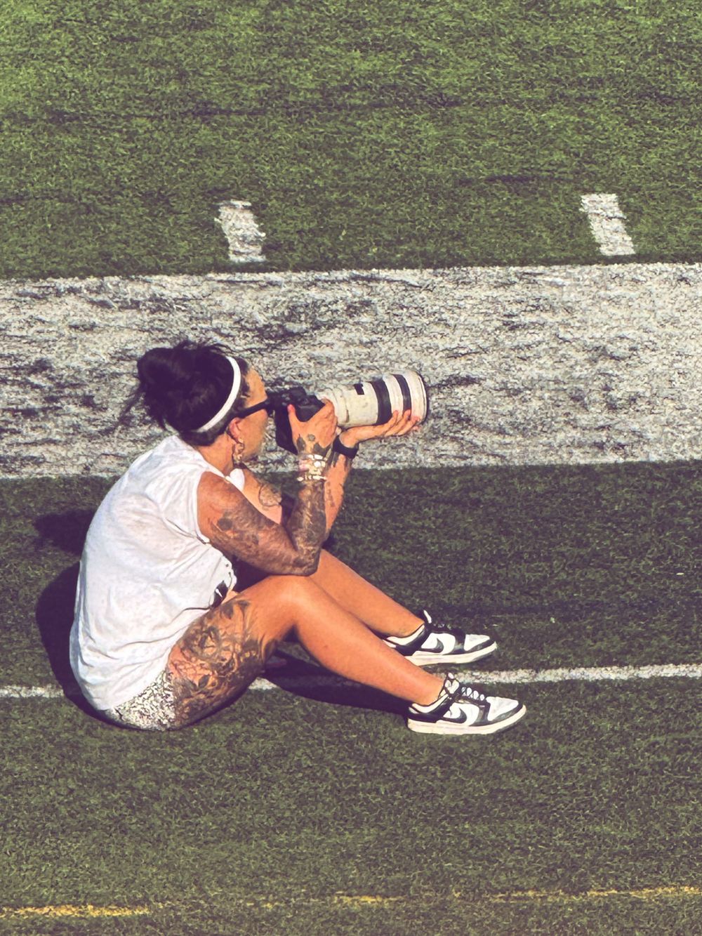 Tattooed photographer sitting on football field sideline, using professional camera with telephoto lens, wearing white outfit.