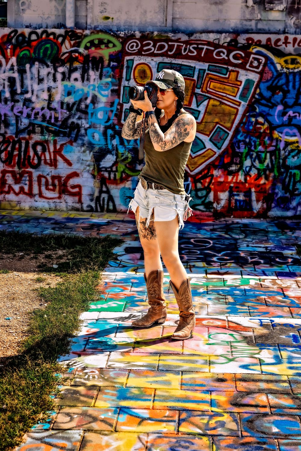 Tattooed woman in denim shorts and cowboy boots photographs graffiti-covered walls on colorful painted ground.