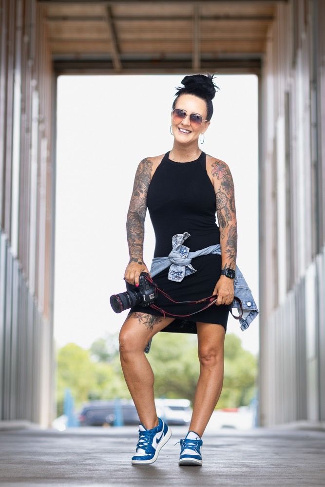 Tattooed woman in black dress and blue sneakers holding a camera, smiling outdoors between columns.