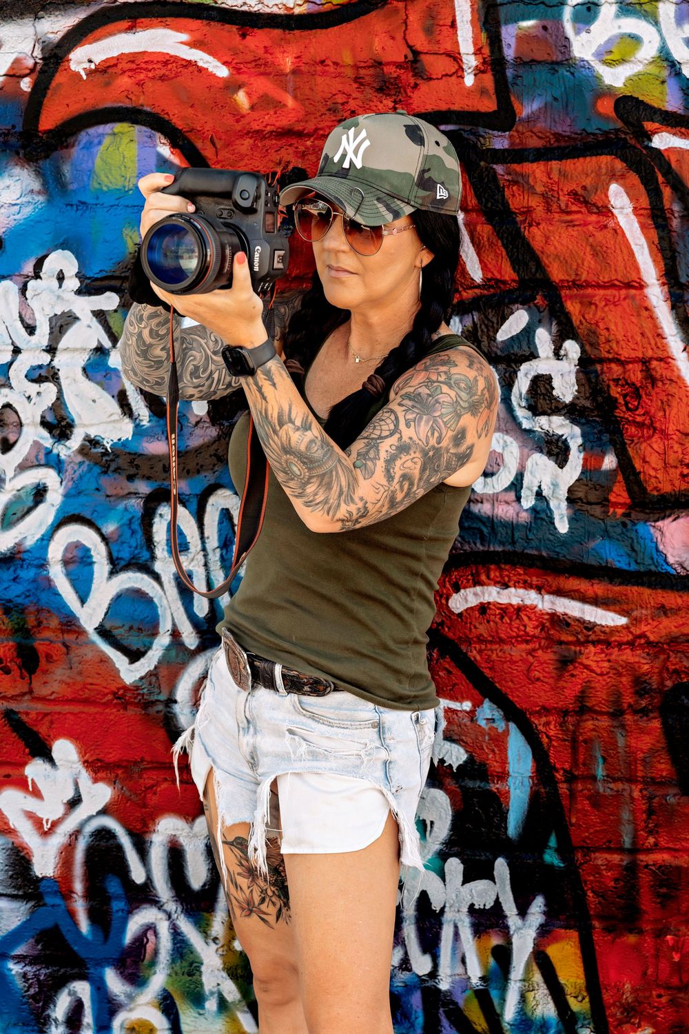 Tattooed woman in NY cap and sunglasses holding a DSLR camera in front of a colorful graffiti wall.