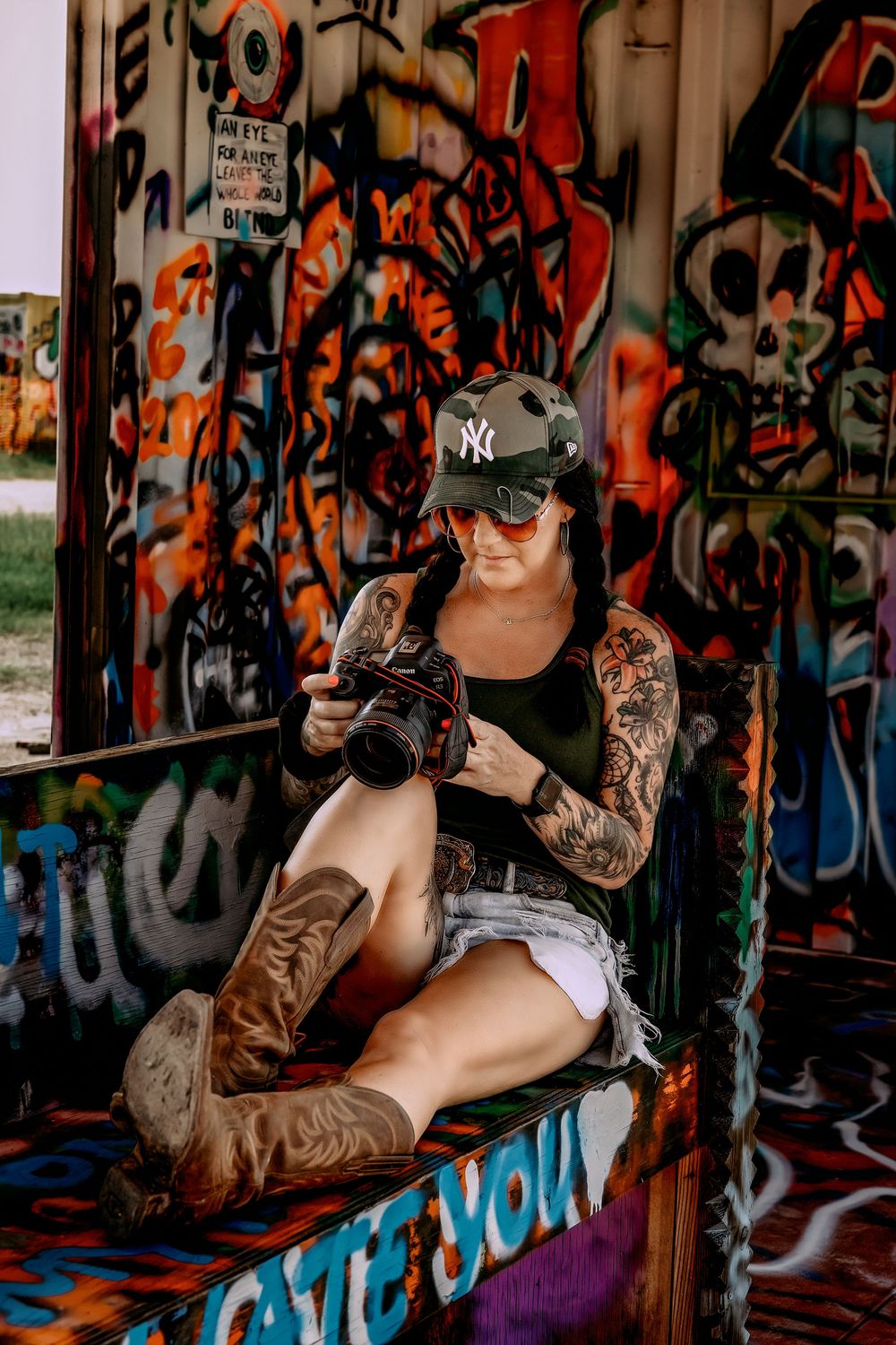 Tattooed woman in NY cap and denim shorts reviewing camera photos against vibrant graffiti wall backdrop.