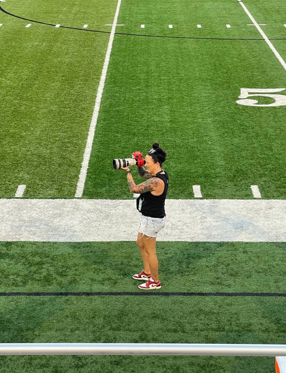 Female photographer with Canon camera on NFL football field sideline, wearing black top and white shorts.