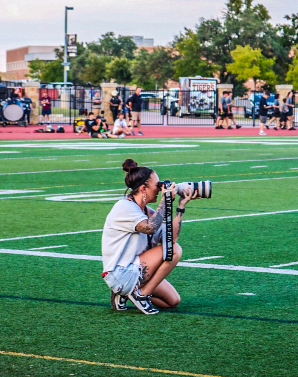 A female photographer kneeling on a green football field, capturing shots with a professional camera and telephoto lens.