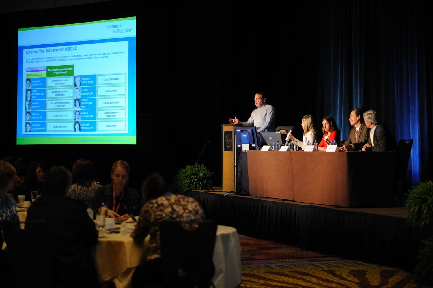 Panel of speakers at a professional conference presentation with projected slides in a large banquet hall.