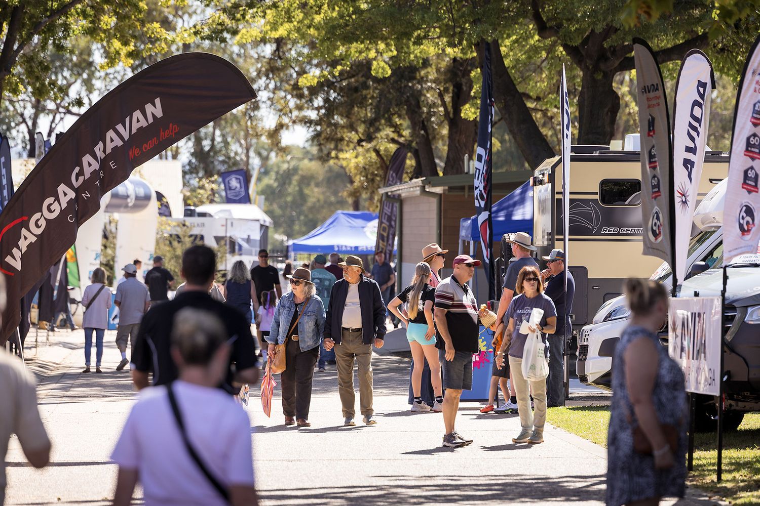 Crowds browse the Wodonga Caravan outdoor show with flags, RVs, and exhibitor stalls under sunny skies.