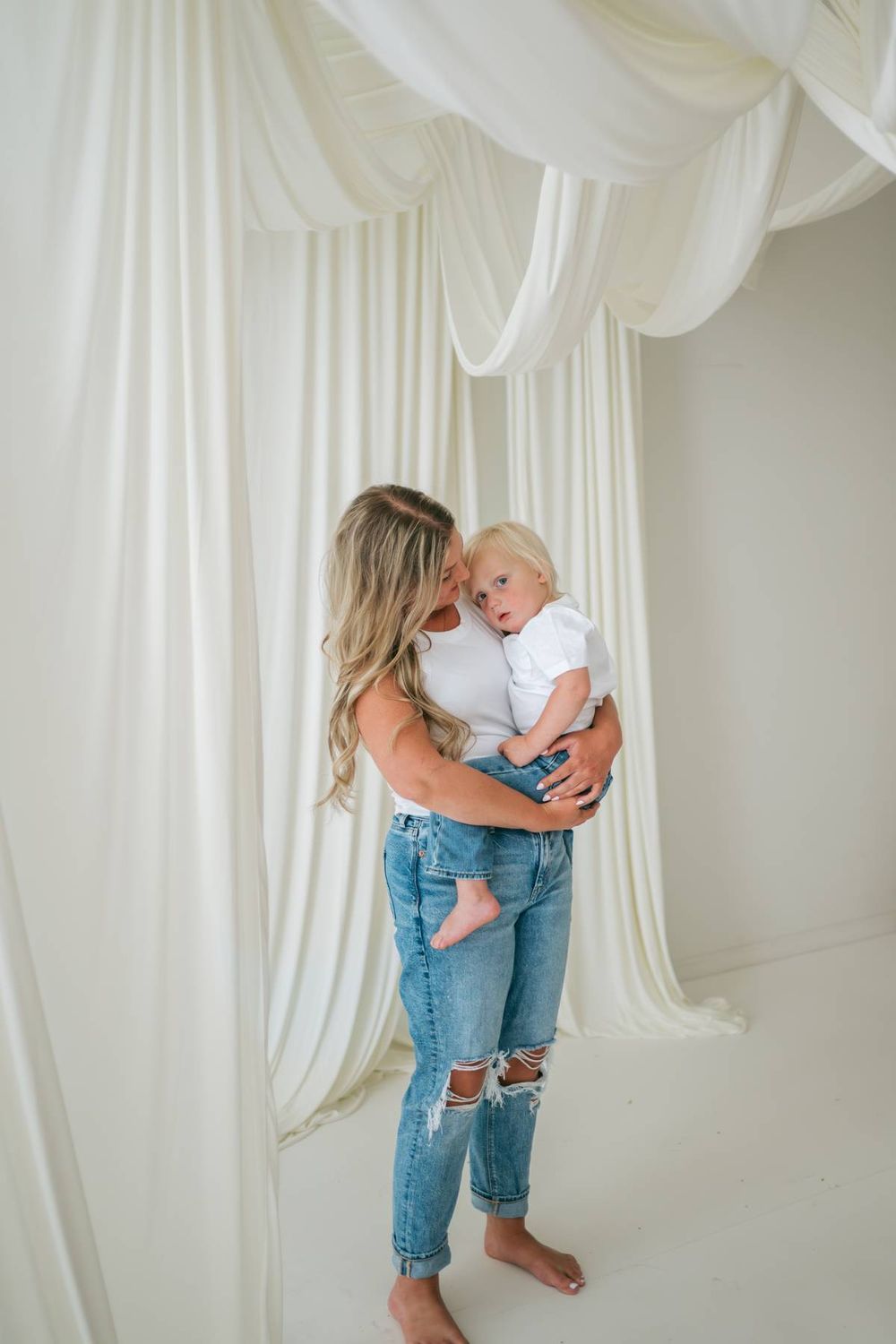 Blonde mother in jeans and white top holding toddler son in a white draped studio in North Carolina