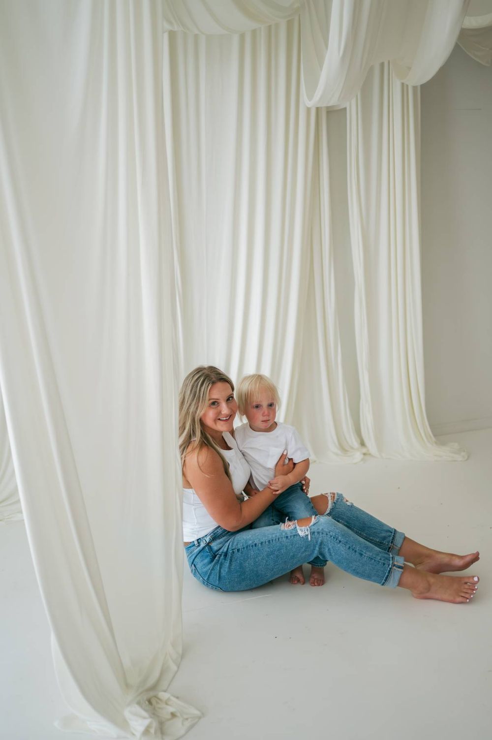 Smiling mother sitting on floor holding blonde toddler surrounded by flowing white curtains in a studio Greensboro NC