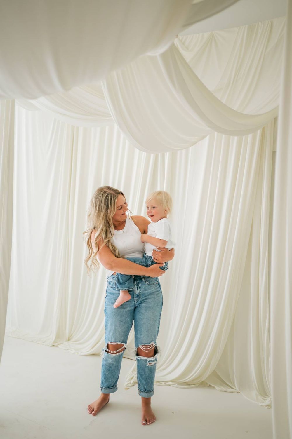 Mother and blonde toddler sharing a playful moment in a bright white fabric draped studio in North Carolina