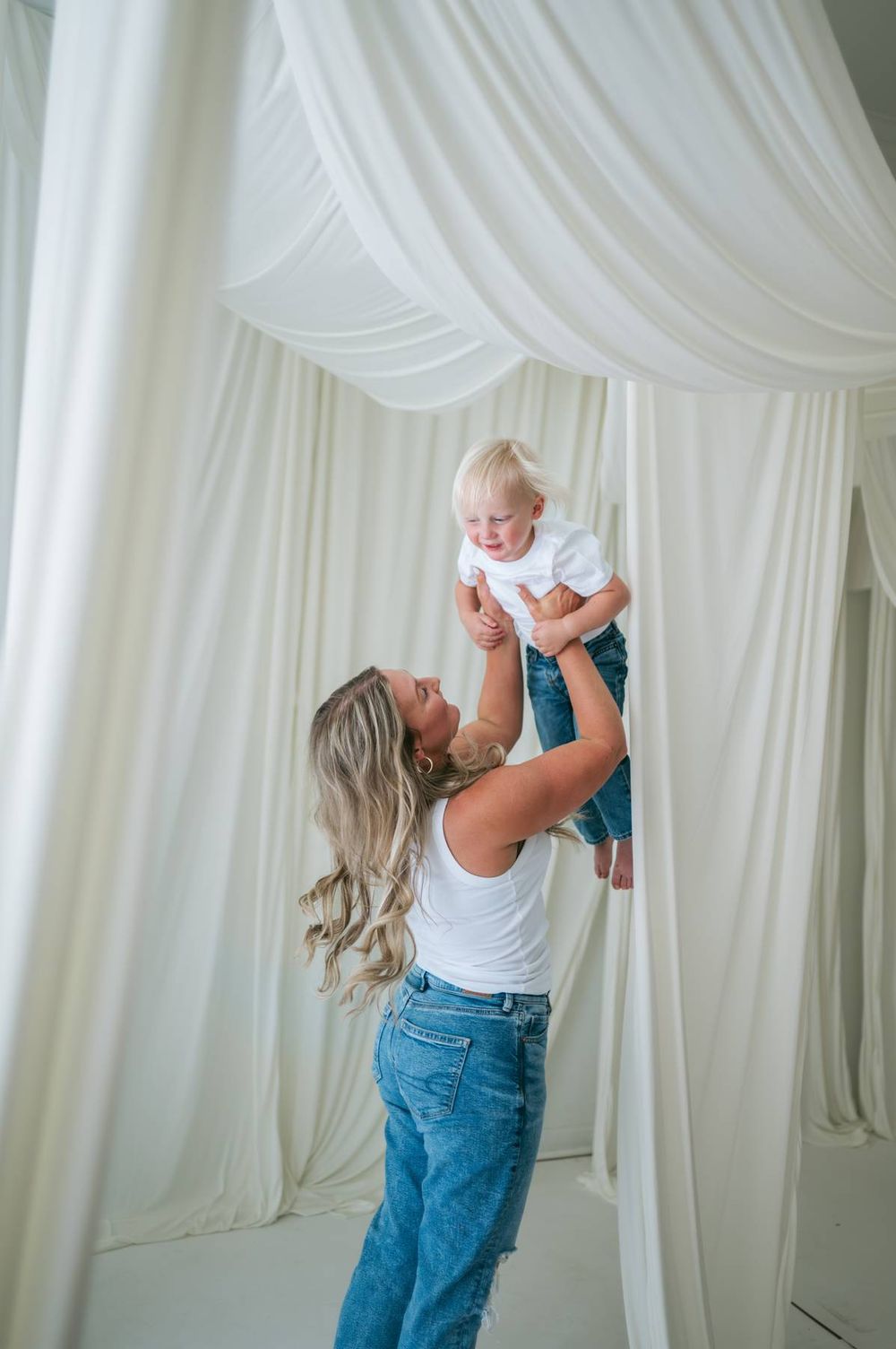 Mother lifting her blonde toddler in the air inside a white draped studio, both wearing matching jeans and white tops North Carolina